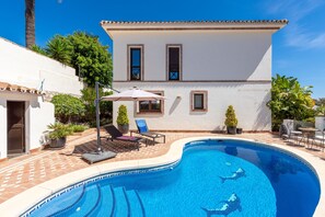 A swimming pool with lounge chairs and umbrellas, a white building with windows, and potted plants.