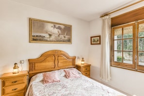 A bedroom with a wooden headboard, a bed with floral bedding, two bedside tables with lamps, a painting on the wall, and a window with curtains.