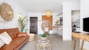 Ein Wohnzimmer mit braunem Sofa, einem runden Kaffeetisch aus Weidwerk mit einer Pflanze, einem Essbereich mit einem hölzernen Stuhl und einer Küche mit weißen Schränken.