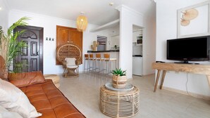 Ein Wohnzimmer mit einer Couch, einem runden Korbtisch mit Pflanze, einem Fernseher auf einem Holzständer und einer Küche im Hintergrund.