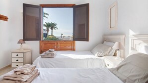 Ein Schlafzimmer mit einem Bett, einem Nachttisch, einer Lampe, einem Fenster mit geöffneten Rollläden und Blick auf Palmen und einen Strand.