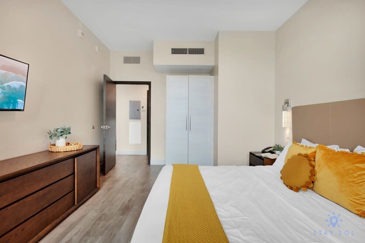 Una habitación de hotel con cama, cómoda de madera, armario, televisor y puerta que da a otra habitación.