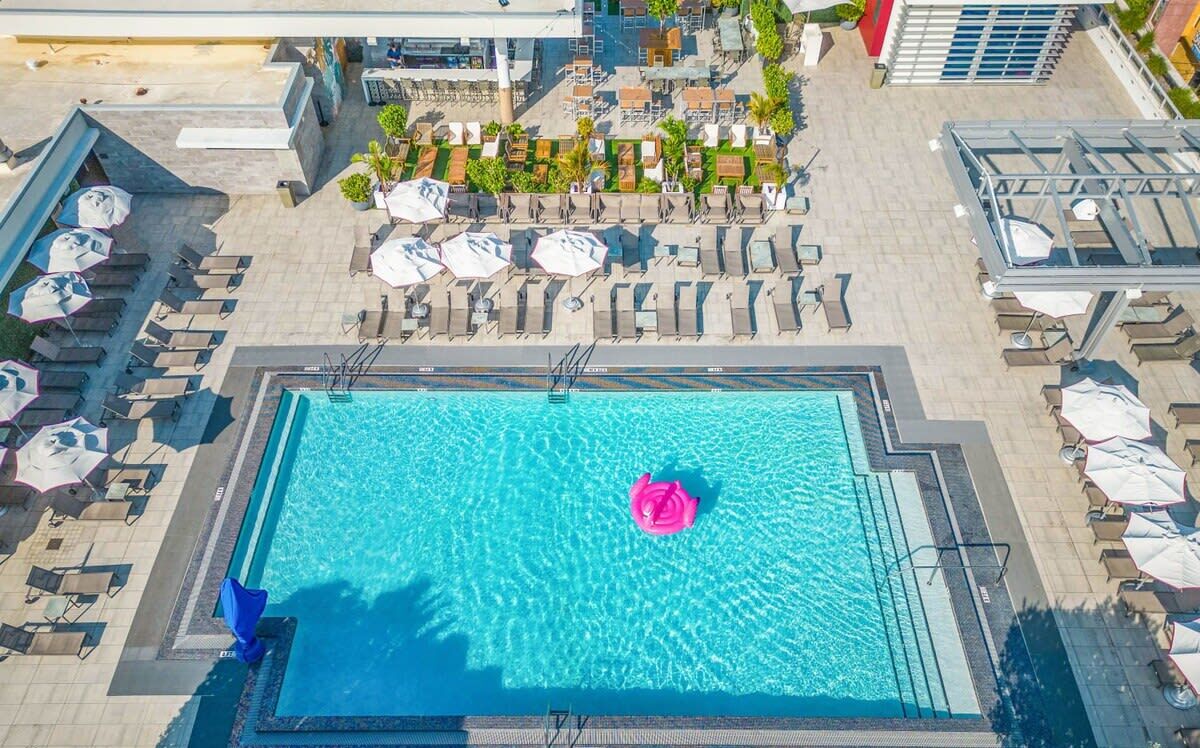 Eine Vogelperspektive auf einen Swimmingpool mit Liegestühlen und Sonnenschirmen, umgeben von einer angelegten Grünfläche mit Bäumen und einem Gebäude im Hintergrund.