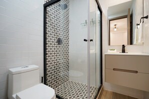 Un baño moderno con cabina de ducha de vidrio, paredes cubiertas de azulejos blancos y un mueble de tocador color beige con espejo.