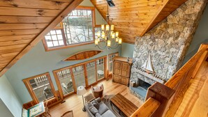 A spacious loft-style living area with a stone fireplace, wooden ceiling beams, and large windows offering a view of the outdoors. - 5000 SF Winnipesaukee Luxury For 14 With 455 Feet Waterfront (Moultonborough)