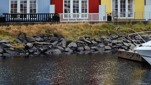 Three colorful houses with white railings and a rocky shoreline in the foreground. - New Boat Houses (Leirvik)