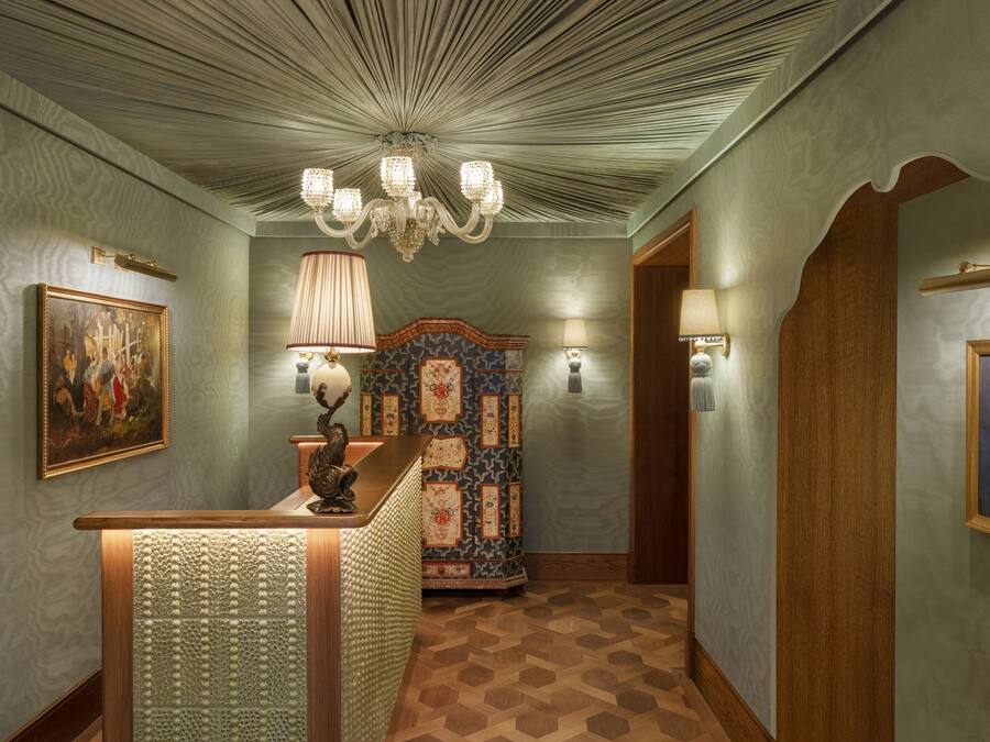 A hallway with patterned wallpaper, a wooden reception desk, a chandelier, and a decorative wall panel.