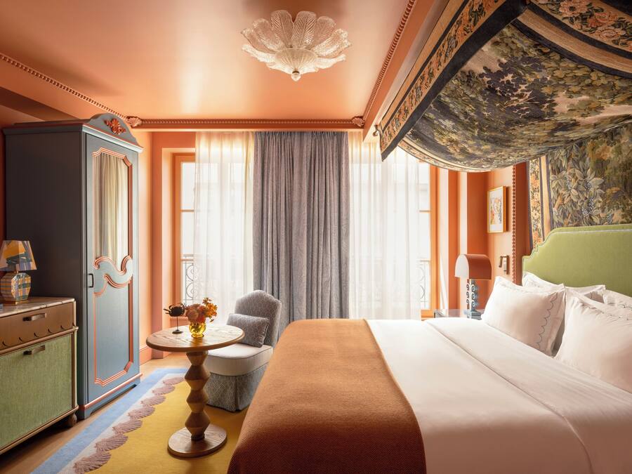 A hotel room with a large bed, a green headboard, a round table with a vase of flowers, a blue wardrobe, and a decorative ceiling.