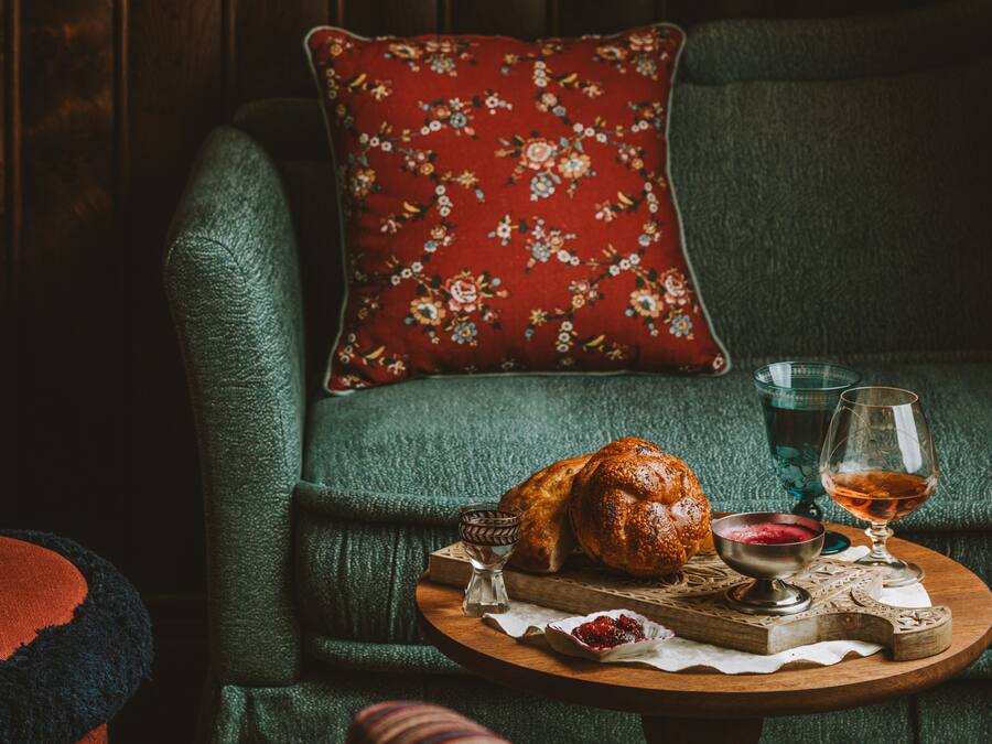 Een gezellige zithoek met een groene bank, een rood kussen met bloemenprint, een houten dienblad met brood, een glas wijn en een klein potje jam.