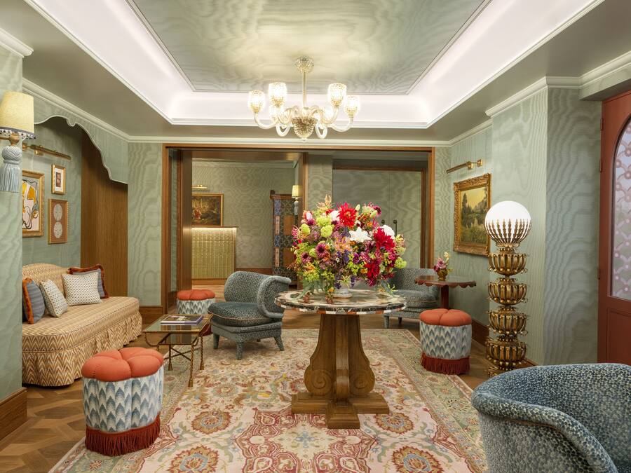 A spacious living room with a central round table, comfortable seating, and a large mirror reflecting the room.