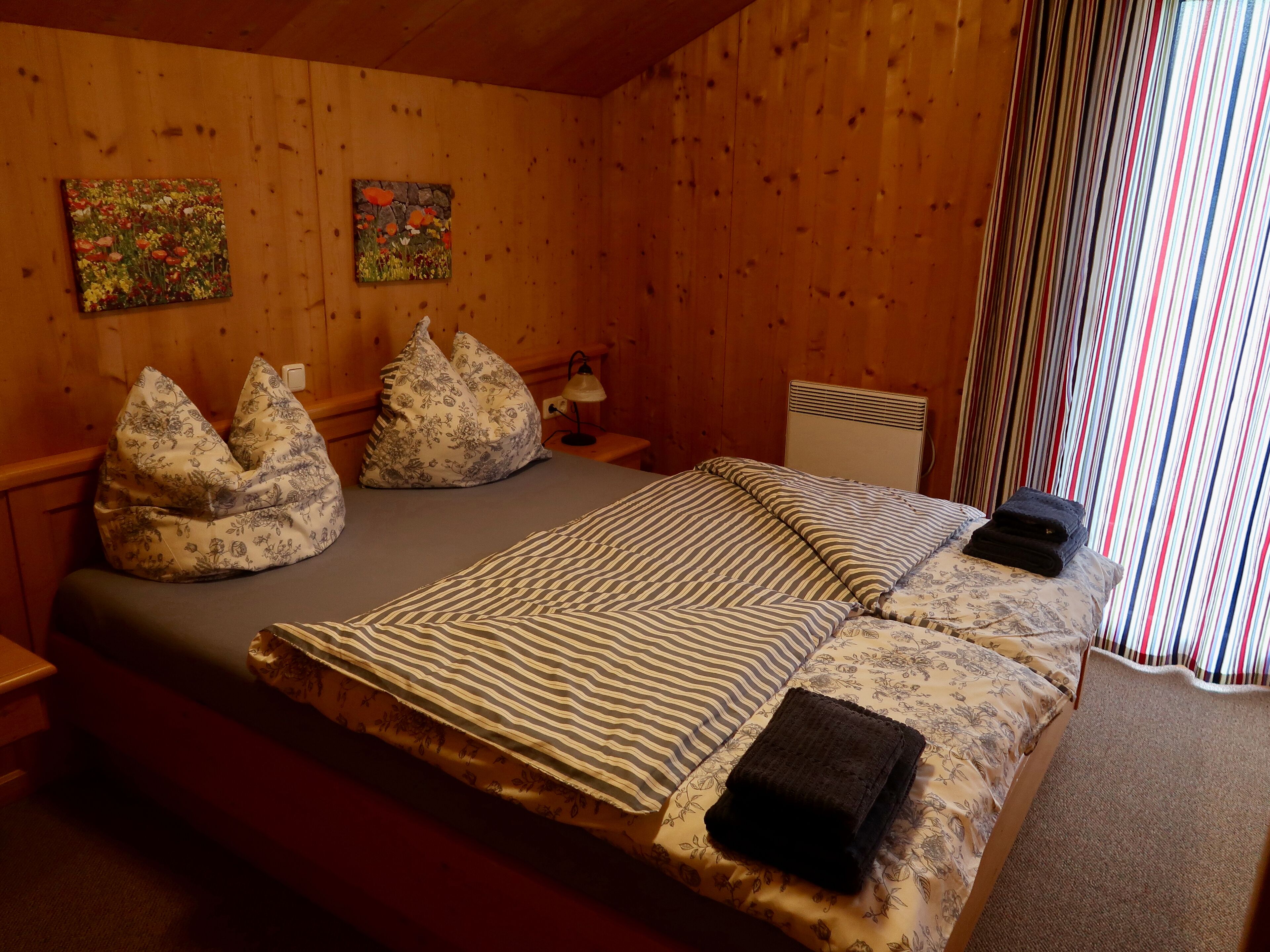A wooden-paneled room with a bed, two bean bags, a nightstand, a lamp, and two paintings on the wall.
