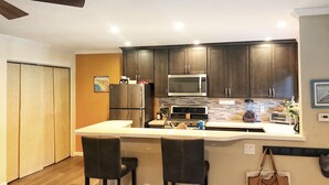 Una cocina moderna con armarios oscuros, un refrigerador de acero inoxidable, un microondas y un horno empotrado. Hay dos taburetes de bar y una encimera con una tetera y un tostador.