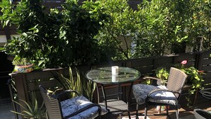 Un patio exterior con sillas de mimbre y una mesa de vidrio, rodeado de plantas en macetas y una cerca de madera.