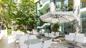 An outdoor cafe with white tables and chairs, a large umbrella, and a glass building in the background. - Hotel Ali d'Oro (Rimini)