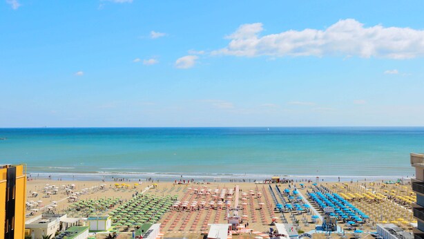 A beach with rows of umbrellas and sunbeds, a clear blue sky, and the ocean in the background. - Hotel Ali d'Oro (Rimini)