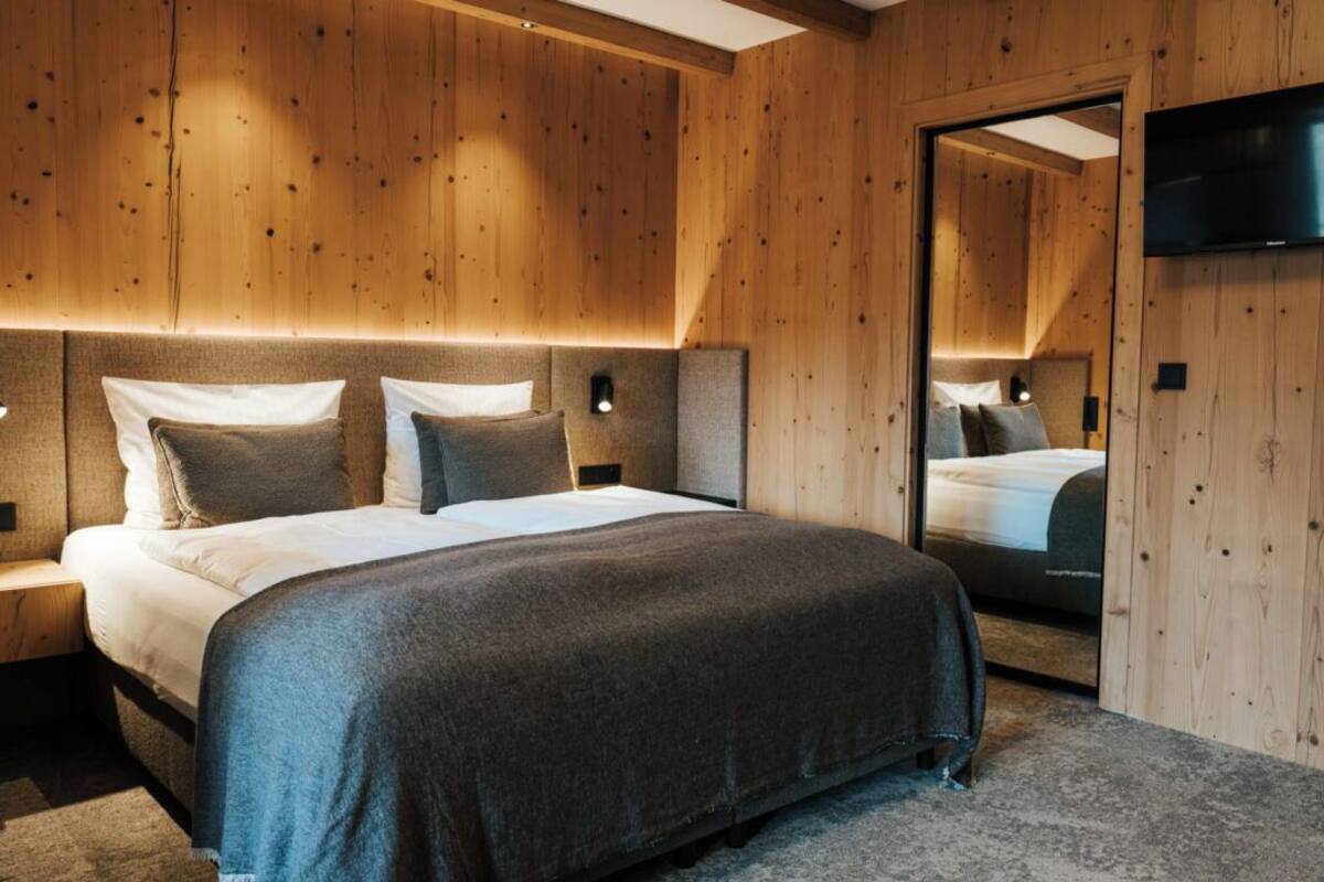 A modern bedroom with a wooden panel wall, a large bed with a grey blanket, bedside lamps, and a TV mounted on the wall.
