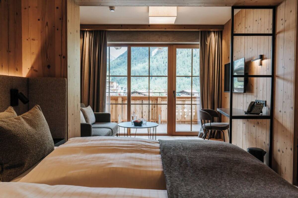 A hotel room with a large bed, a small table with chairs, a TV mounted on the wall, and a view of mountains through the window.
