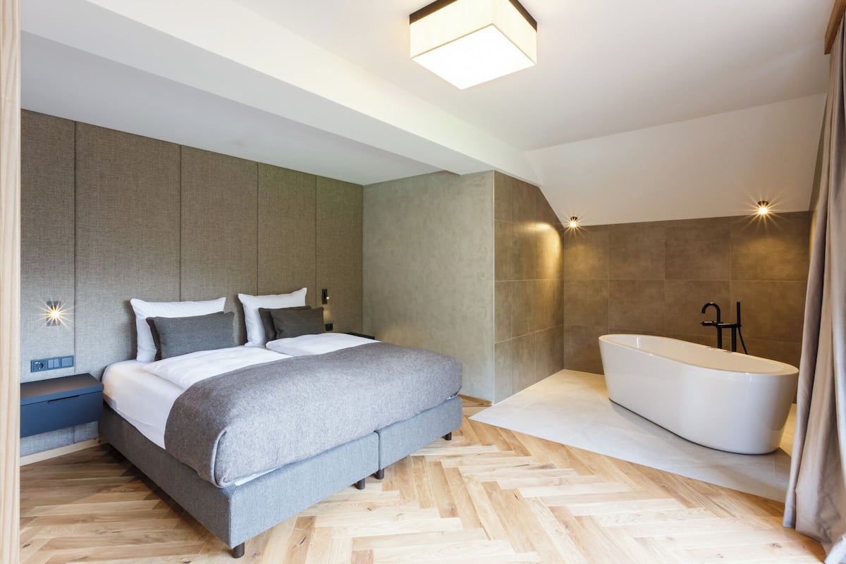 A modern bedroom with a large bed, a built-in bench, a bathtub, and a herringbone wood floor.