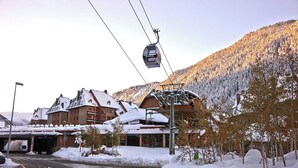 A ski lift with a cable car moving over a snowy landscape, a building with a ski lift on its roof, and a road with snowbanks on either side. - Val de Ruda Luxe 16 by FeelFree Rentals (Baqueira)