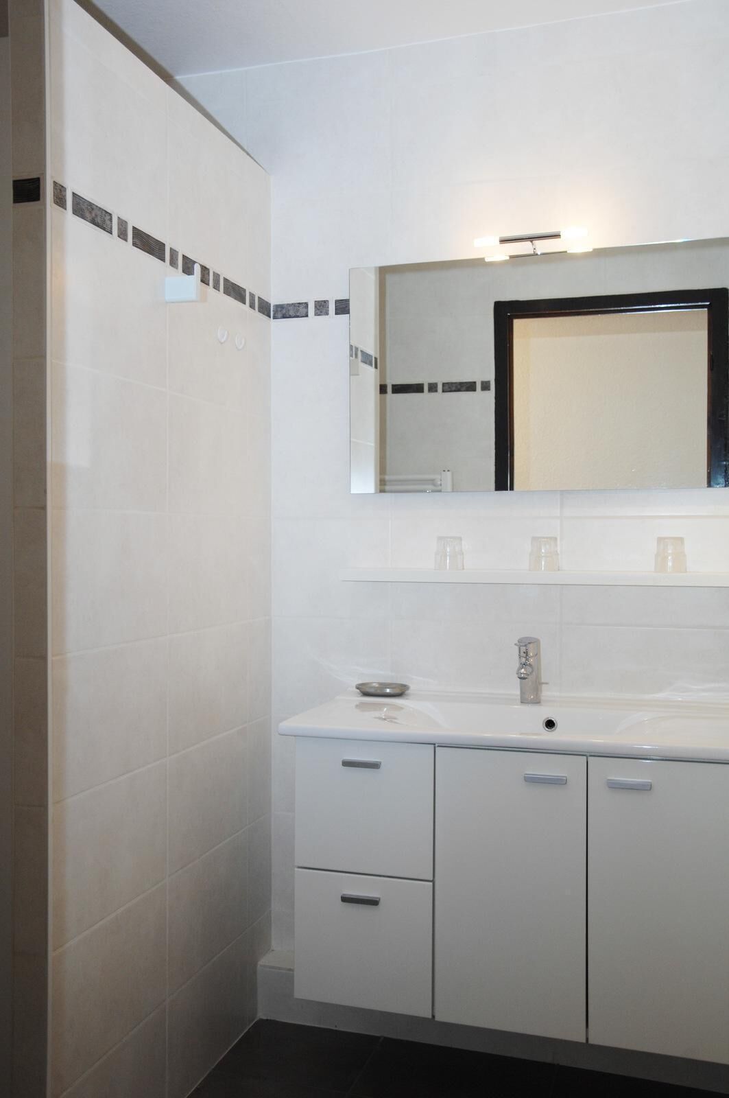 A modern bathroom with white tiles, a large mirror, a sink with a faucet, and a cabinet with drawers.