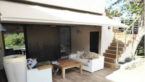 Un espace de vie extérieur moderne, doté d’une terrasse en bois, de mobilier blanc et d’un escalier menant à un niveau supérieur.
