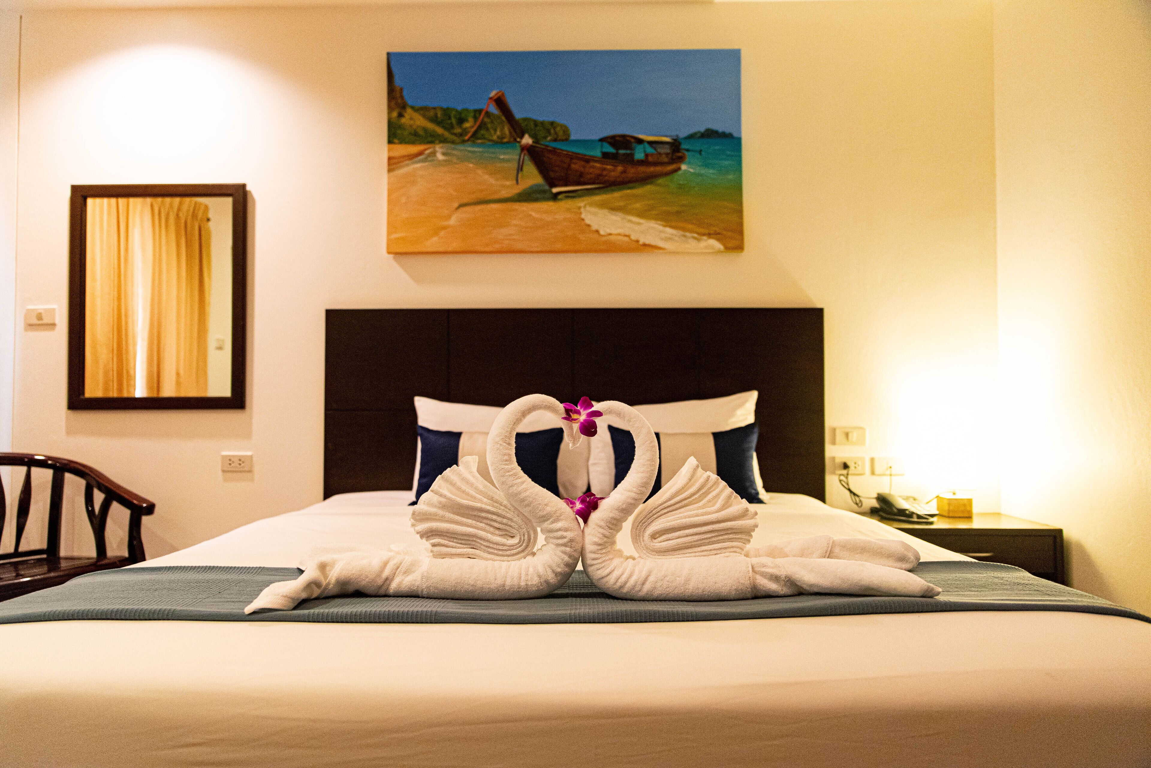 A hotel room with a bed, a painting of a boat on the beach, and a pair of swan-shaped towels on the bed.