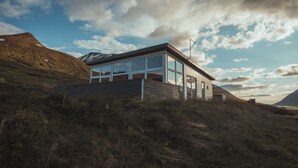 Een modern huis met grote ramen, gelegen op een grasheuvel met een berg op de achtergrond.