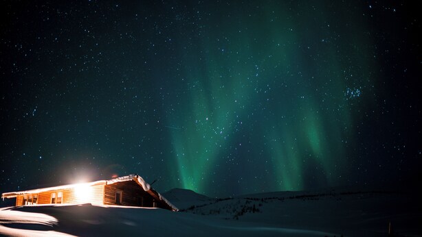 A snow-covered landscape with a wooden cabin illuminated by lights, under a starry night sky with aurora borealis. - Hidden Cabin with outdoor hot tub (Ólafsfjordur)