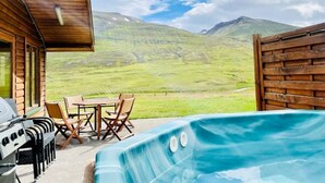 A wooden cabin with a hot tub and barbecue grill, overlooking a grassy landscape and mountains. - Hidden Cabin with outdoor hot tub (Ólafsfjordur)