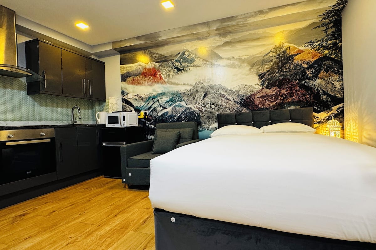 A modern hotel room with a large bed, a kitchenette, and a mural of a mountainous landscape.