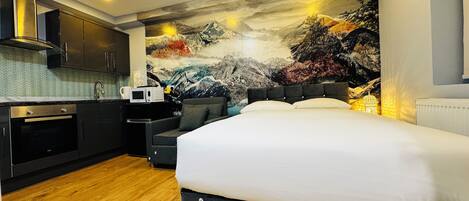 A modern hotel room with a large bed, a kitchenette, and a mural of a mountainous landscape.