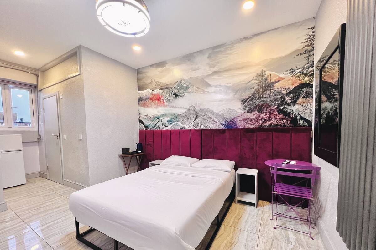 A hotel room with a bed, a small desk, a chair, a TV, and a mural on the wall.