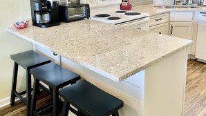 Una cocina con armarios blancos, encimera de granito, estufa, microondas, cafetera y dos taburetes de bar.