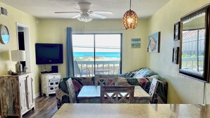 Una habitación con cocina, una mesa de comedor con sillas, un ventilador de techo, televisión y vista a la playa.