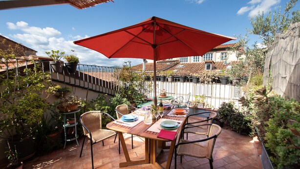 An outdoor patio with a wooden table set for dining, surrounded by wicker chairs, and a red umbrella providing shade. - Residenza al Pavone Luxury with Terrace (Lucca)