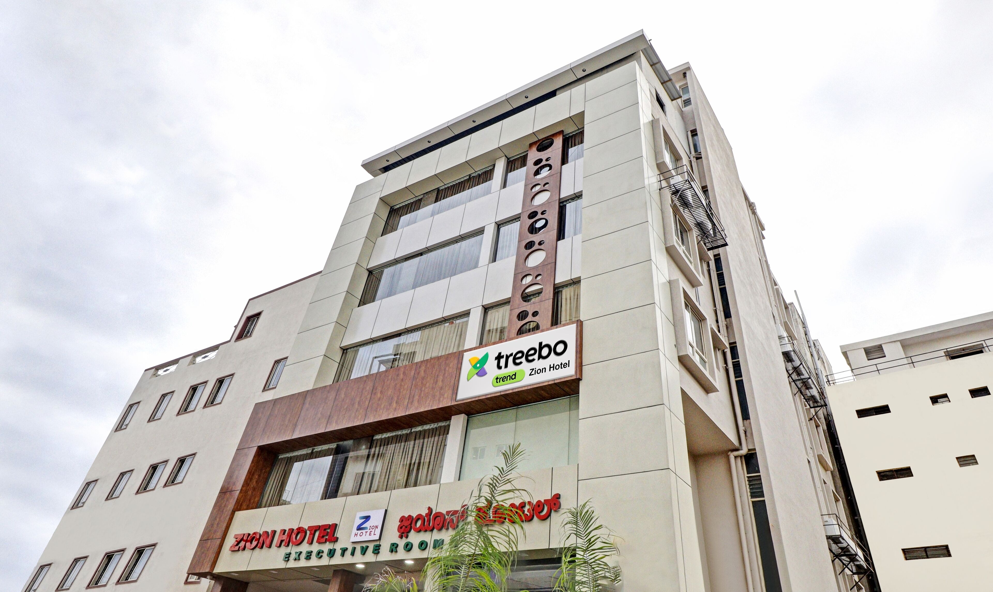 Un hotel de varios pisos con un diseño moderno, que destaca un cartel prominente de 'Zion Hotel' y el logo de 'Treebo' en la fachada.