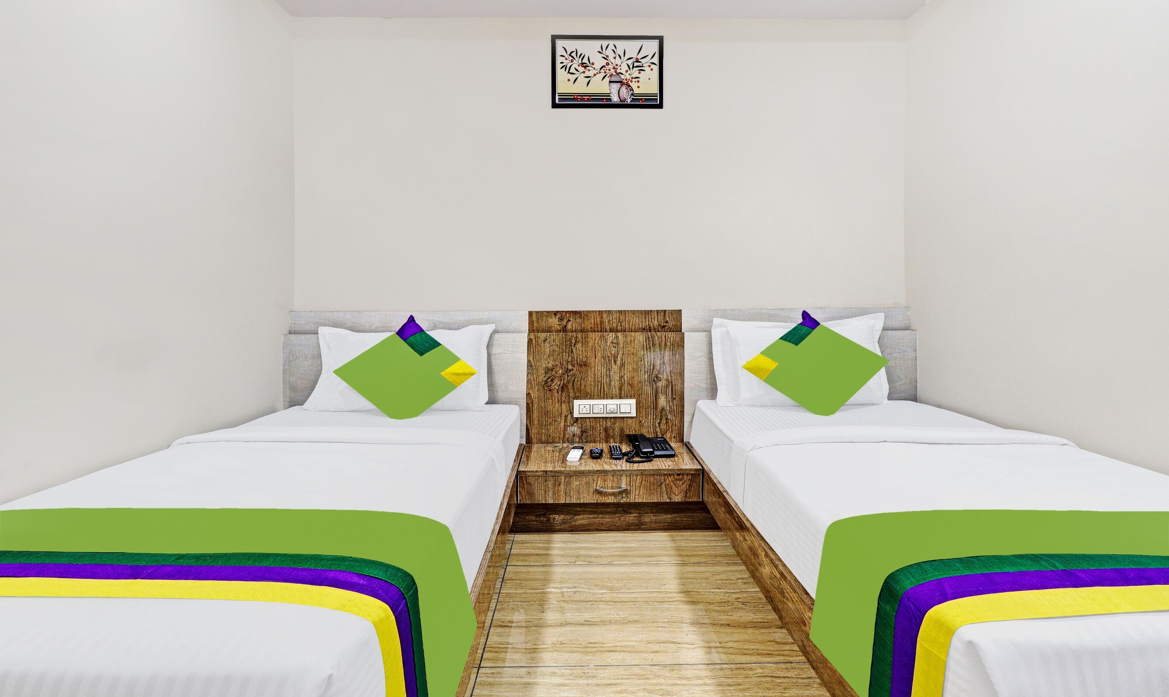 A hotel room with two beds, a wooden headboard, a small table with a phone and remote, and a wall-mounted light switch.