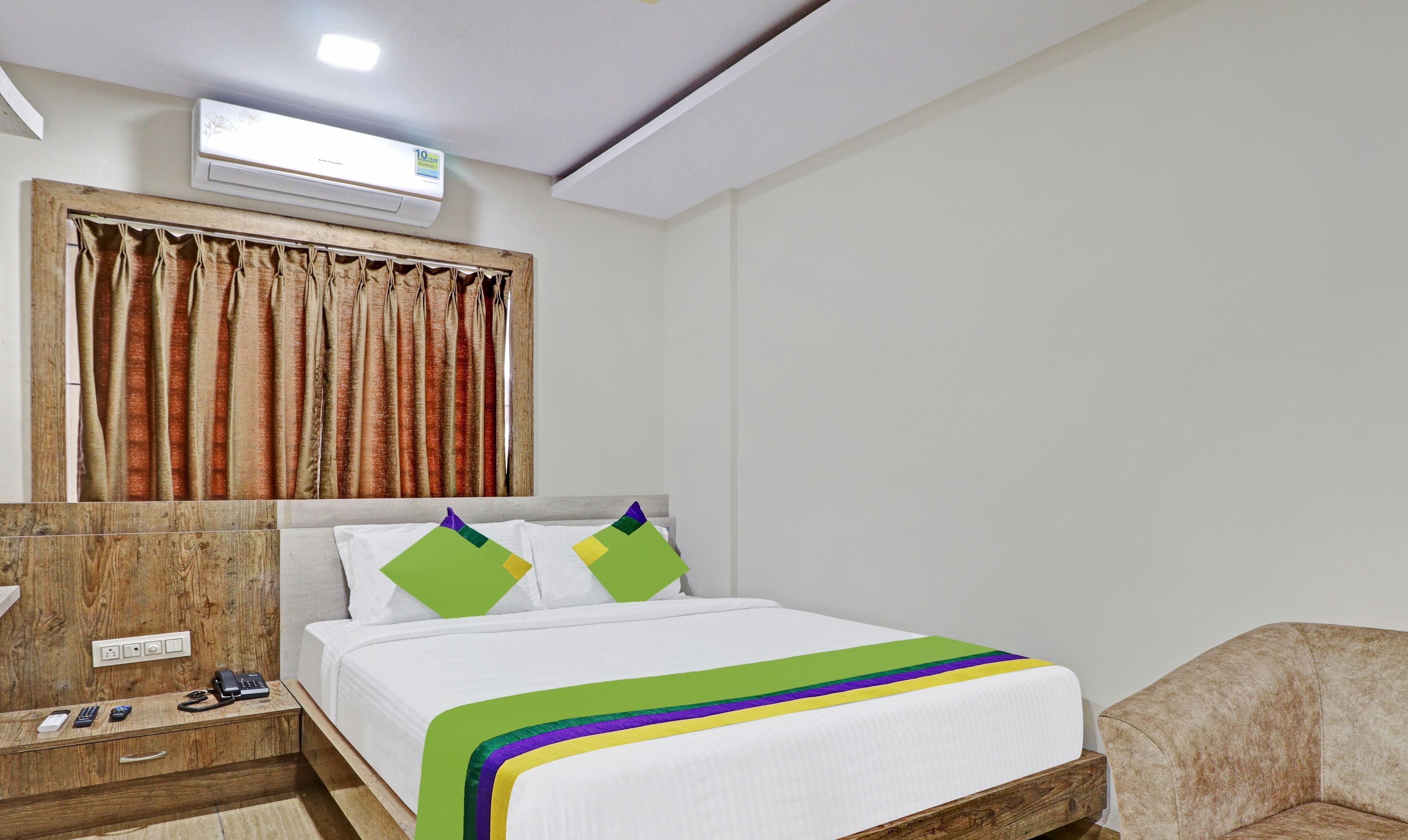 A hotel room with a bed, a wooden nightstand with a phone and remote controls, a wooden headboard, and a window with curtains.