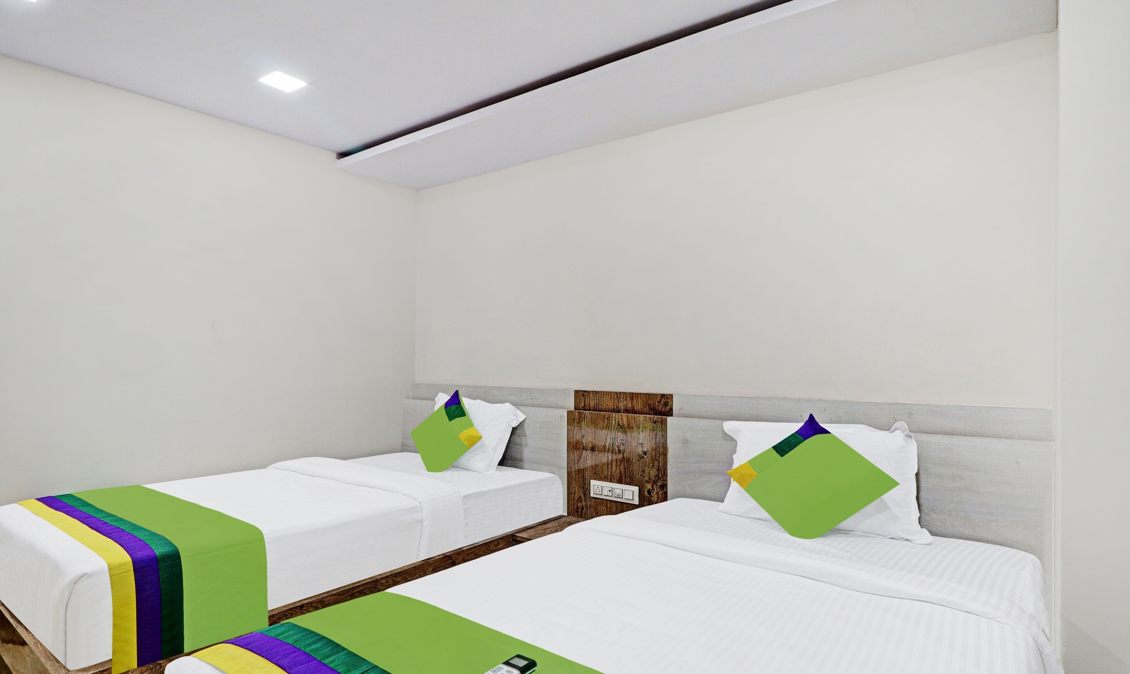 A hotel room with two single beds, each with a white mattress and a colorful bedspread. The room has a wooden headboard and a white wall.