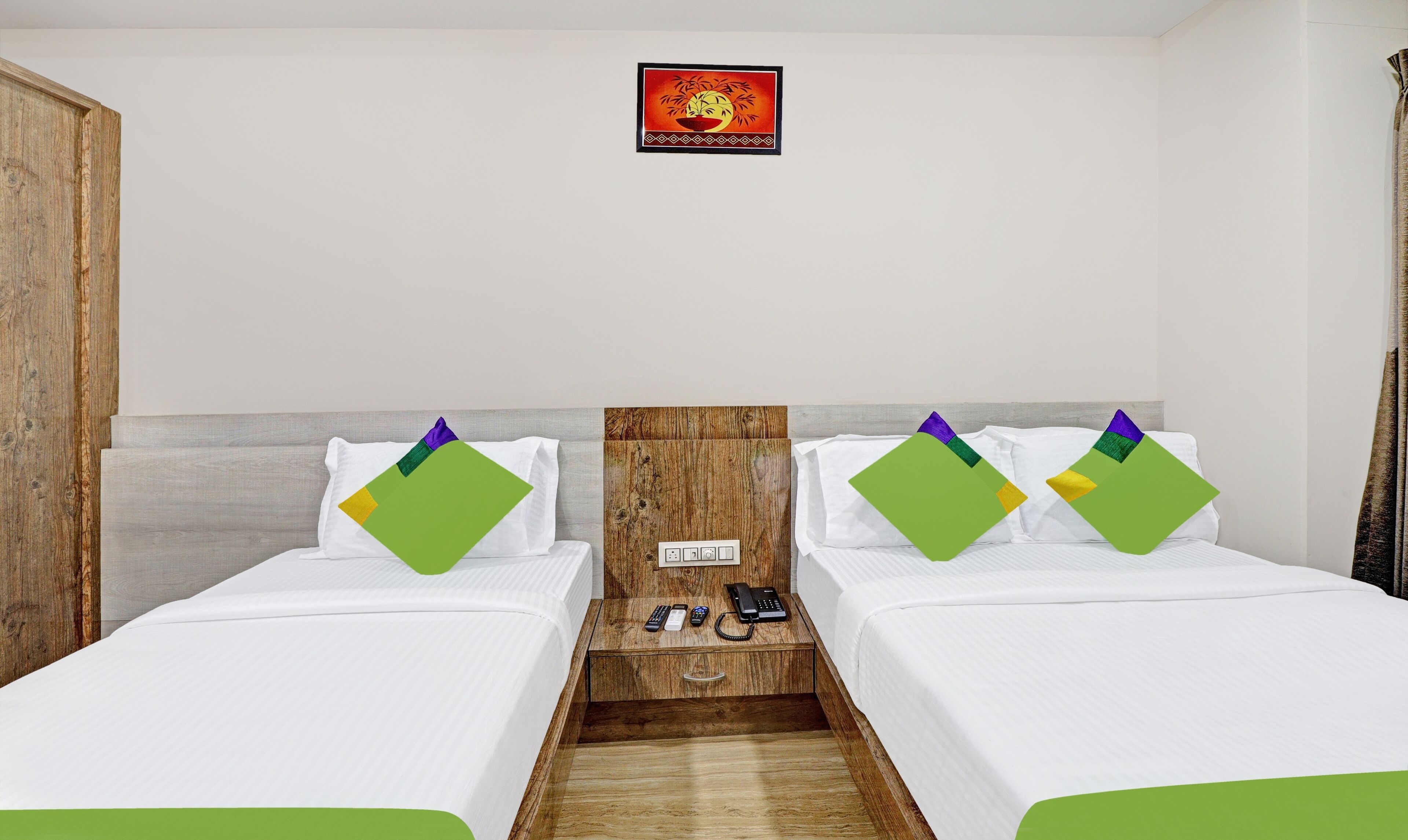 A hotel room with two single beds, a wooden headboard, a small wooden desk with a telephone and remote controls, and a wall-mounted sign above the bed.