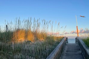 Ein hölzerner Steg führt zu einem Strand mit hohem Gras und blauem Himmel.