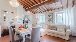 A spacious living room with a wooden ceiling, a dining area with a white table and chairs, and a white sofa with decorative pillows. - Anna Farmhouse Apartment in Wine Resort in Lucca (Monte San quirico)