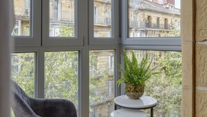 Una habitación con ventanas amplias, una silla gris, una mesita con una planta y vistas a edificios.