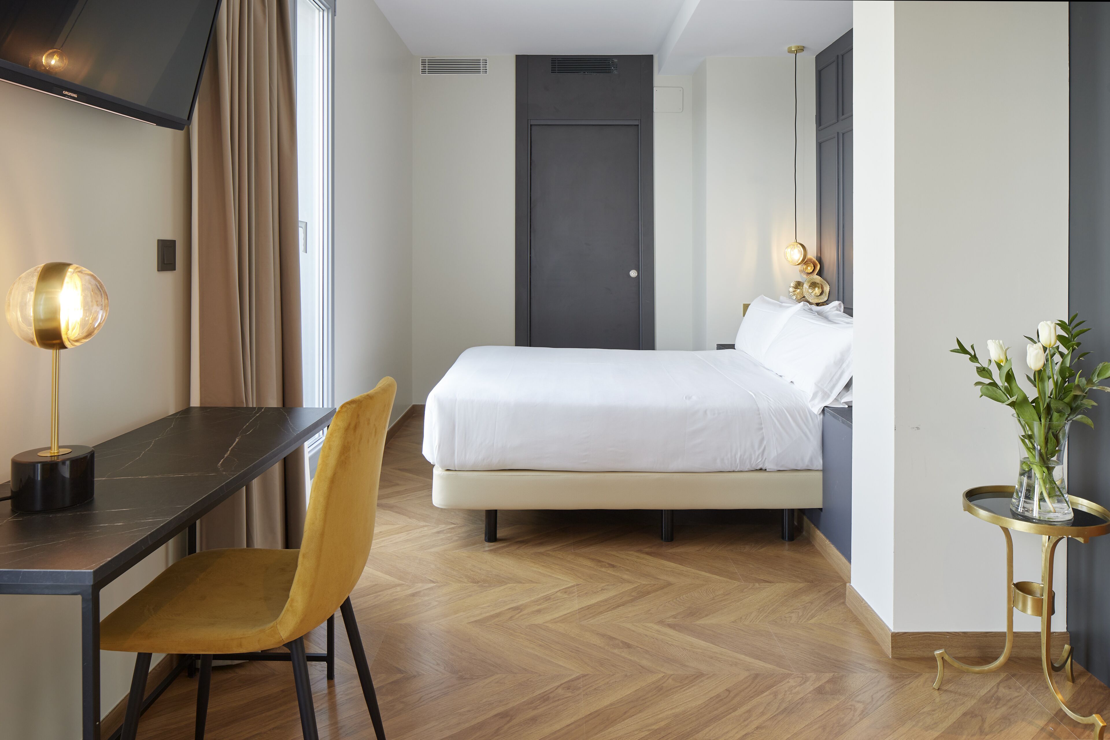 A hotel room with a bed, a desk, a chair, a TV, and a plant.