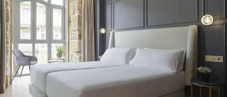 A double bed with white linens in a room with a stone wall and a window with curtains.