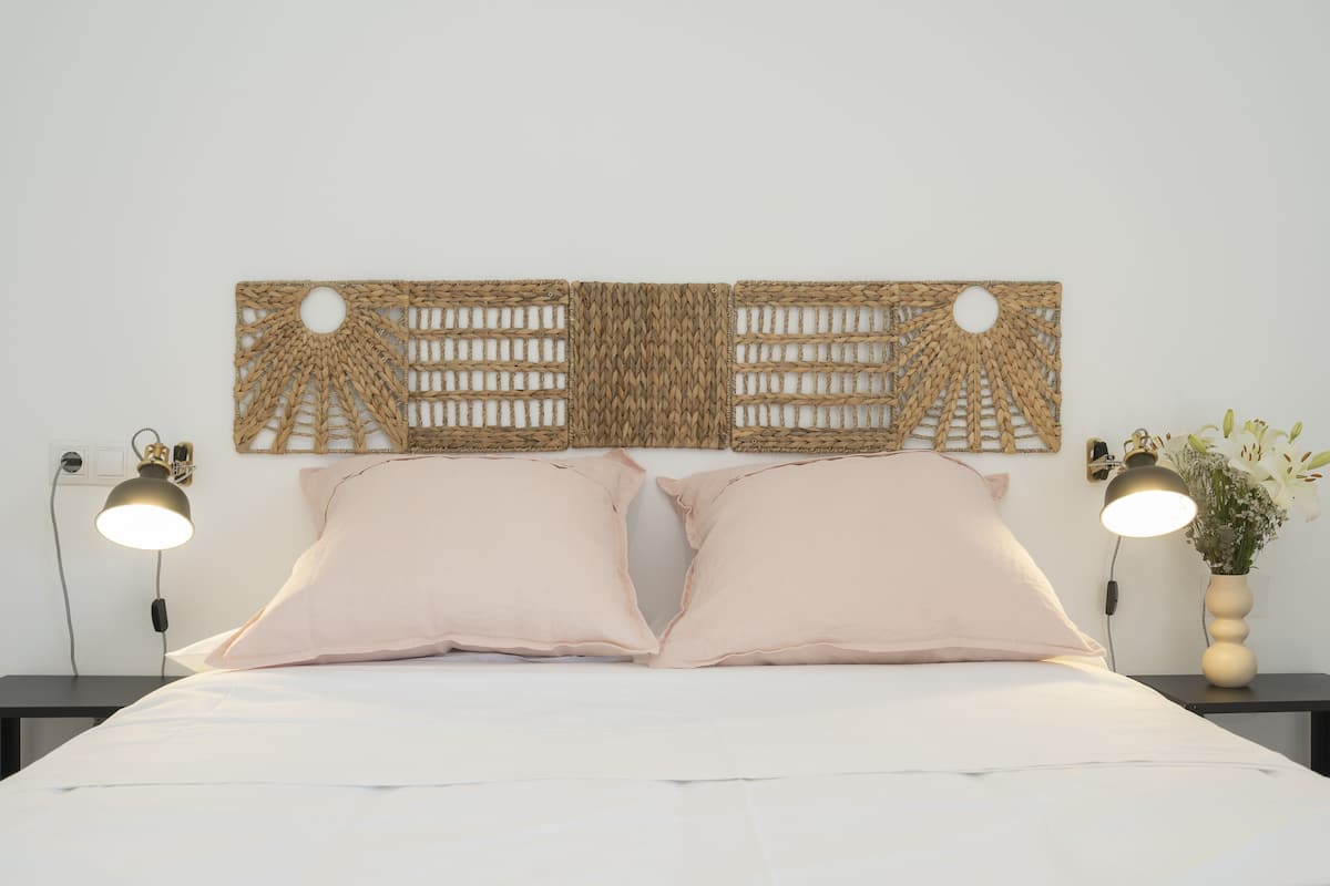 Una cama con dos almohadas, un aplique en la pared y una mesita de noche con una lámpara y un florero.