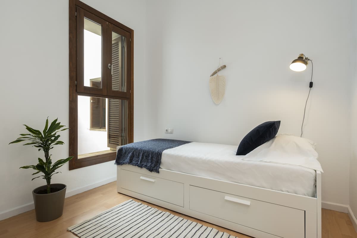 Un dormitorio con cama, planta en maceta, ventana con persianas y lámpara de pared.