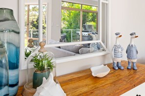 A room with a large window, a wooden table, decorative items, and a view of greenery outside. - Bonito near Bagnalls Beach Corlette (Corlette)