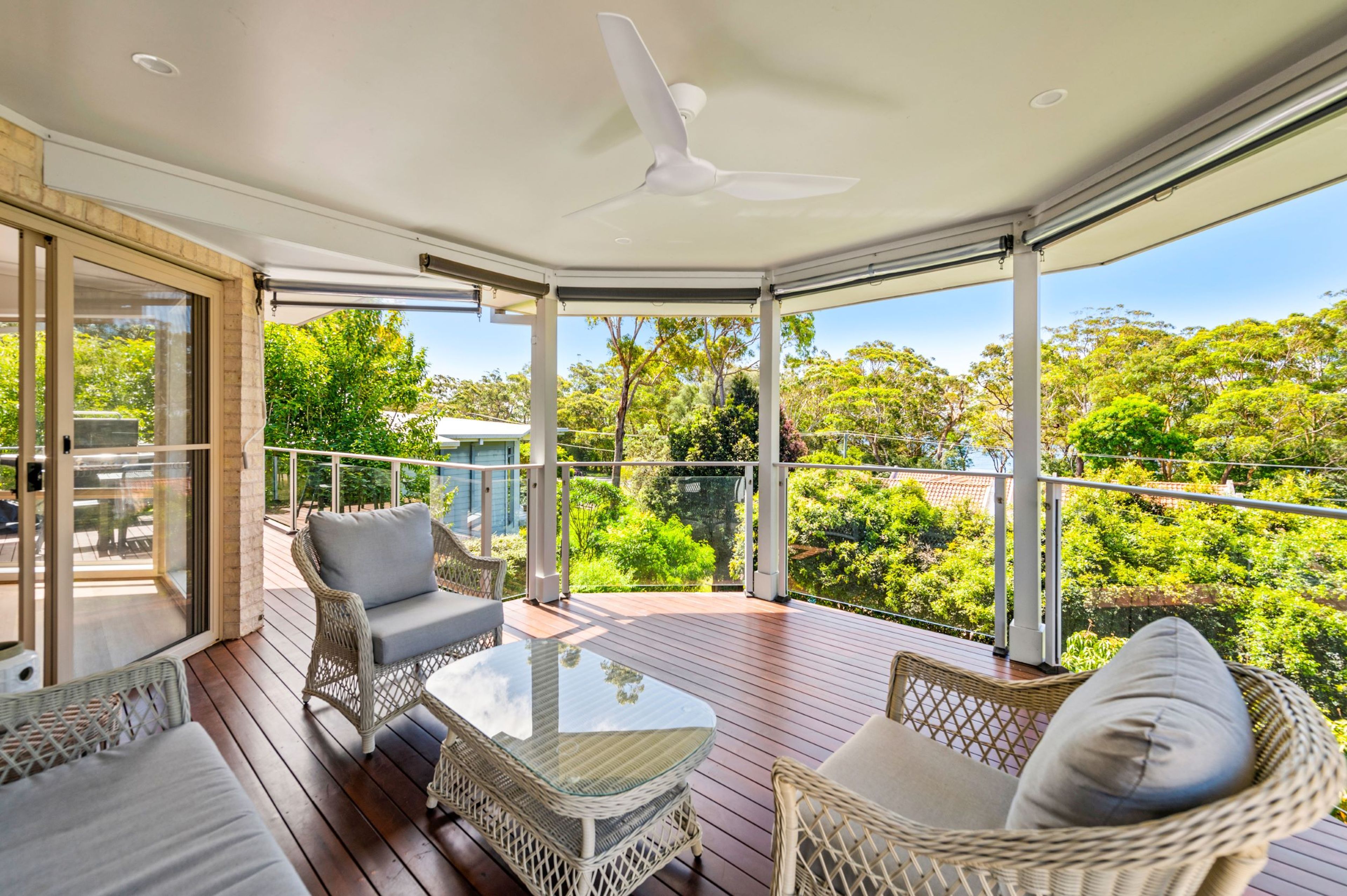 A covered outdoor area with wicker furniture, a glass table, and a view of greenery.