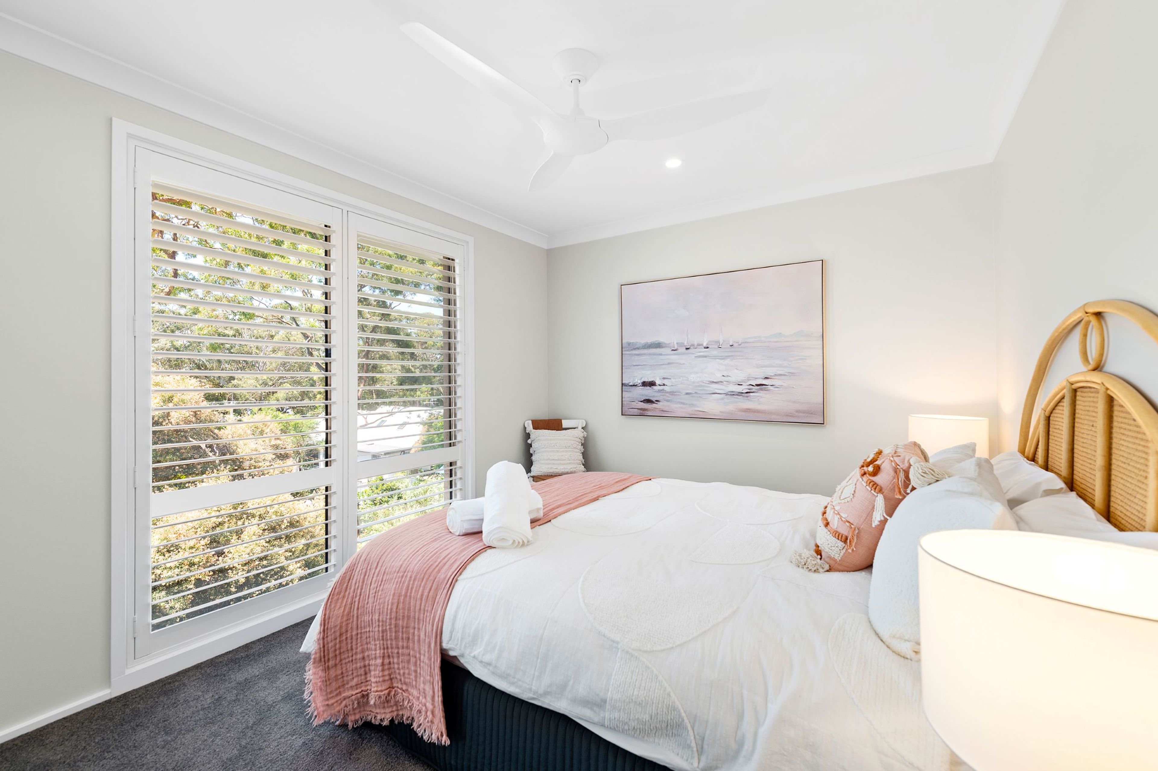 A bedroom with a bed, a ceiling fan, a window with blinds, a painting on the wall, and a bedside table with a lamp.